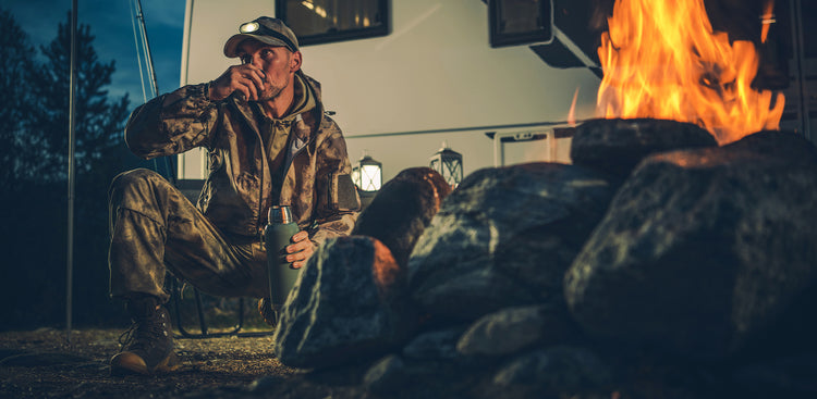 A man sitting in a chair while camping wearing full camouflage and drinking out of a thermos bottle in front of a fire.  