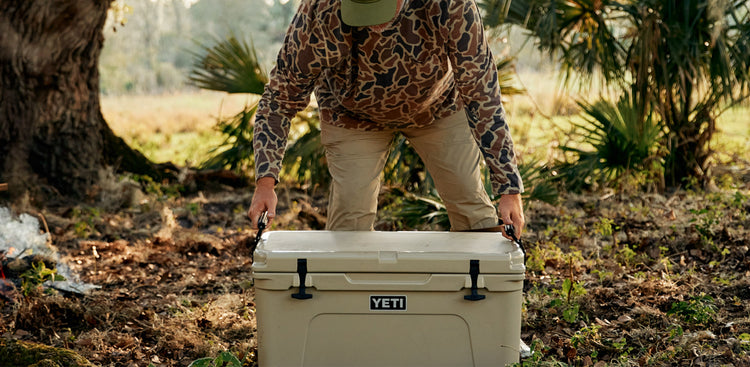 A man wearing an old school camo pullover and khaki pants, picking up a tan YETI cooler in an open area in front of a tree